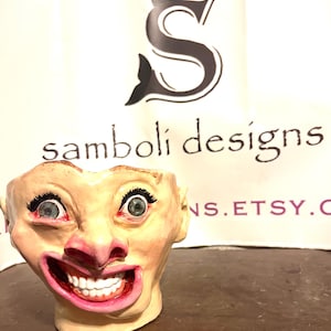 May include: A ceramic face planter with a wide, exaggerated smile and open mouth, featuring detailed teeth and pink lips. The face has green eyes with long eyelashes and is painted in flesh tones. The planter is a unique decorative item.