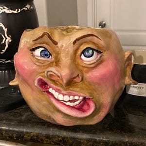 Könnte beinhalten: Ein Keramik-Pflanztopf in Form eines menschlichen Gesichts mit einem breiten, zahnigen Grinsen. Das Gesicht ist mit realistischen Details bemalt, darunter blaue Augen, rosige Wangen und eine leicht krumme Nase.
