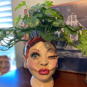 Pode incluir: Um vaso de cerâmica em forma de cabeça de mulher com cabelo ruivo, piscando um olho e com blush rosa. O vaso tem uma planta verde que cresce no topo.