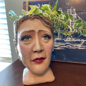 May include: A ceramic planter shaped like a woman's head with a leafy green plant growing out of the top. The planter has realistic features, including painted eyes, lips, and eyebrows.