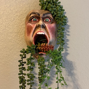 Peut inclure: Sculpture murale d'un visage hurlant avec des yeux verts et une bouche ouverte. Le visage est peint avec des tons rouges et bruns. Des vignes vertes et feuillues tombent du haut et de la bouche du visage, avec quelques petites fleurs oranges.