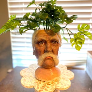 May include: A unique planter shaped like an elderly man's head, with a white mustache and a sad expression. The planter holds vibrant green artificial plants. The planter sits on a decorative, scalloped base with a carved design.