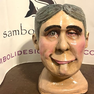 May include: A painted ceramic bust of a person with gray hair and a smiling expression. The bust has a light skin tone with pink cheeks and lips. The text "sambolidesic" is visible in the background.