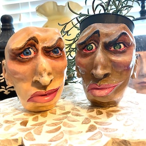 May include: Two ceramic face planters with expressive features. One is light-skinned with blue eyes, the other is dark-skinned with green eyes. Both have pink lips and are on a decorative, mosaic-tiled stand. One holds greenery.