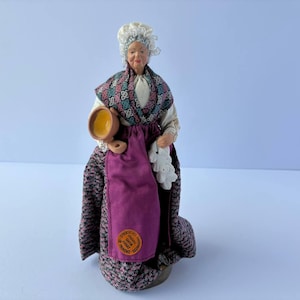 May include: A ceramic figurine of a woman wearing a white cap, a purple skirt, and a patterned shawl. She is holding a brown pot in her right hand and a white cloth in her left hand. The figurine is standing on a wooden base.