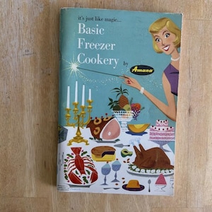 May include: Vintage cookbook cover titled "Basic Freezer Cookery by Amana." The cover features an illustration of a woman with blonde hair, pointing a wand at a table laden with food, including a turkey, cake, and lobster.