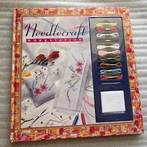 May include: A book titled "Needlecraft Workstation" with a colorful floral design on the cover. The book features a variety of embroidery projects, including a small pillow, a bookmark, and a piece of fabric with a needle and thread. The book also includes a set of colorful embroidery floss.