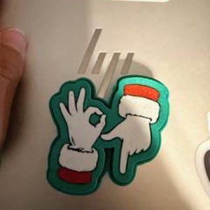 May include: A green and white shoe charm featuring two hands making hand gestures. One hand forms an 'okay' sign, and the other points downward. The hands have white gloves with red and white cuffs. The charm is on a gray surface.