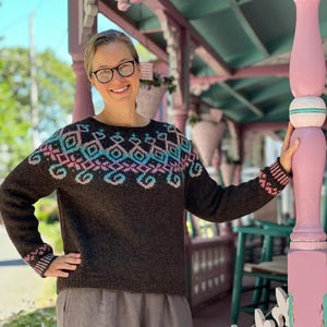 May include: A woman wearing a dark brown sweater with a geometric pattern in teal and pink. The sweater has matching cuffs and is paired with gray linen pants. The background includes a porch with pink and white accents.