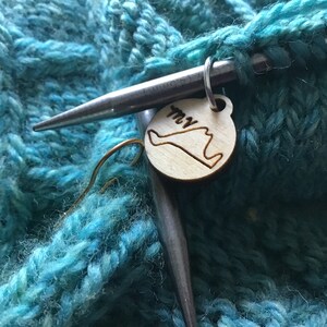 May include: A close-up of a teal knitted fabric with a wooden stitch marker in the shape of the state of Long Island, New York. The marker is attached to a silver knitting needle.