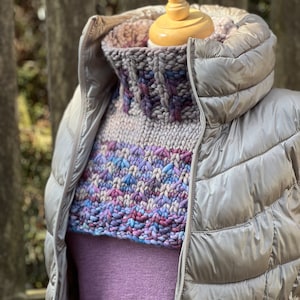 May include: A chunky knit cowl neck sweater in shades of purple, pink, and blue, worn over a purple long-sleeved shirt and a gray puffer jacket.