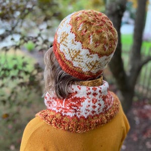 May include: A knitted hat and neck warmer set in a fall color palette of orange, white, and brown. The hat has a geometric pattern and the neck warmer has a snowflake pattern.