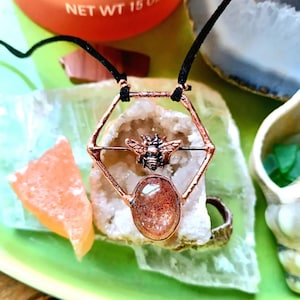 May include: A copper wire hexagon pendant with a sunstone and a small bee charm. The pendant is hanging from a black cord.