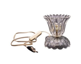 May include: A clear glass table lamp with a fluted shade and a faceted base. The lamp has a white cord with an inline switch and a black plug. The lamp is designed to sit on a table or desk.