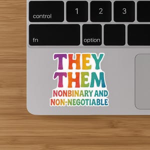May include: A sticker with the words "THEY THEM" in a rainbow gradient font, with the text "NONBINARY AND NON-NEGOTIABLE" below. The sticker is on a laptop, near the keyboard.
