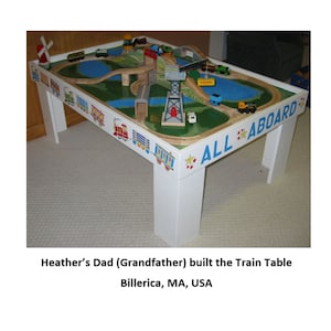 Puede incluir: Una mesa de tren de madera blanca con un diseño de vías de tren pintadas, con un paisaje verde, elementos de agua azules y varios trenes de juguete. El borde de la mesa está decorado con ilustraciones de trenes coloridas y las palabras "ALL ABOARD".