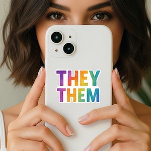 May include: A white smartphone featuring a rectangular sticker on the back. The sticker displays the text THEY THEM in a bold, sans-serif font. The letters feature a vibrant, multi-colored watercolor gradient transitioning from purple to blue.