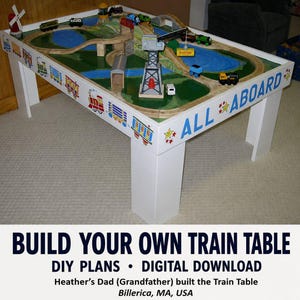 May include: A white wooden train table featuring a painted surface with green landscapes, blue water, and wooden tracks. The sides are decorated with colorful train illustrations and the text ALL ABOARD. It includes a crane, bridge, and toy trains.