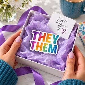 May include: A white gift box filled with purple tissue paper containing a sticker with the text THEY THEM in a watercolor rainbow gradient. A small white card with the text Love You and a heart symbol is tucked inside the box.