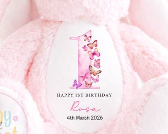 May include: A pink plush bunny with embroidered eyes and nose. The bunny has a number one design with butterflies and the text "HAPPY 1ST BIRTHDAY Rosa 4th March 2026". The text "Newborn Safe: Embroidered Eyes & Nose" is also visible.