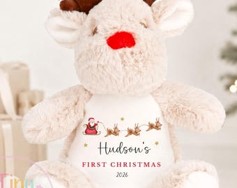 May include: A plush beige reindeer toy with dark brown antlers and a bright red nose. The center of the chest features a white patch printed with a graphic of Santa in a sleigh, the text "Hudson's FIRST CHRISTMAS 2026," and small gold stars.