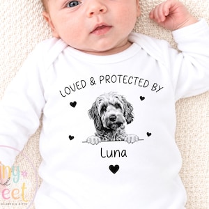 May include: A white long-sleeve baby bodysuit featuring a black graphic of a dog resting its paws on a line. Above the dog, the text reads LOVED & PROTECTED BY, and below the dog, the name Luna is printed, surrounded by small black heart icons.