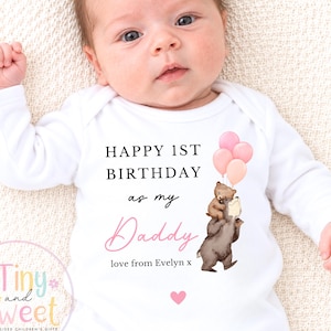 May include: A white long-sleeve baby bodysuit featuring the black text HAPPY 1ST BIRTHDAY as my Daddy, the pink script text Daddy, and the text love from Evelyn x. An illustration shows two brown bears holding three pink balloons and a pink heart.