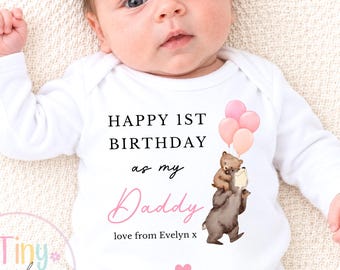Puede incluir: Un body blanco de manga larga para bebé con el texto negro HAPPY 1ST BIRTHDAY as my Daddy, la palabra Daddy en letra cursiva rosa y el texto love from Evelyn x. Una ilustración muestra dos osos pardos con tres globos rosas y un corazón.