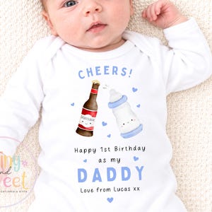 May include: A white long-sleeve baby bodysuit featuring a graphic of a beer bottle and a baby bottle with the text CHEERS! Happy 1st Birthday as my DADDY Love from Lucas xx. The design includes small blue hearts surrounding the two illustrated items.