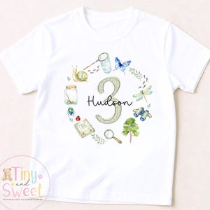 May include: A white short-sleeve t-shirt featuring a large number 3 with the name Hudson printed across it. The design includes a circular arrangement of watercolour illustrations of a snail, butterfly, dragonfly, binoculars, magnifying glass, and jar.