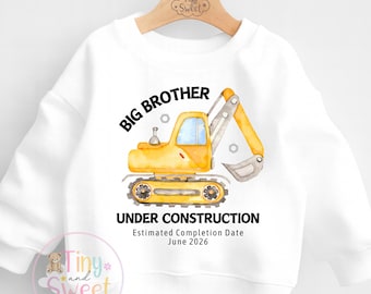 May include: White sweatshirt featuring a yellow excavator with the words "BIG BROTHER" and "UNDER CONSTRUCTION." The estimated completion date is June 2026. The sweatshirt has a crew neck and long sleeves.