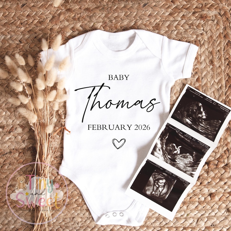 May include: A white baby onesie with the name "Thomas" in elegant script, the month and year "February 2026", and a heart symbol. Next to the onesie are ultrasound images and dried flowers, all on a woven background.