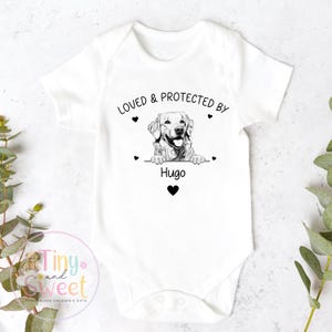 Puede incluir: Un body blanco de manga corta para beb&eacute; con un estampado gr&aacute;fico en negro de un perro golden retriever apoyando sus patas sobre una l&iacute;nea. El texto "LOVED & PROTECTED BY" aparece sobre el perro y "Hugo" debajo, con peque&ntilde;os corazones.