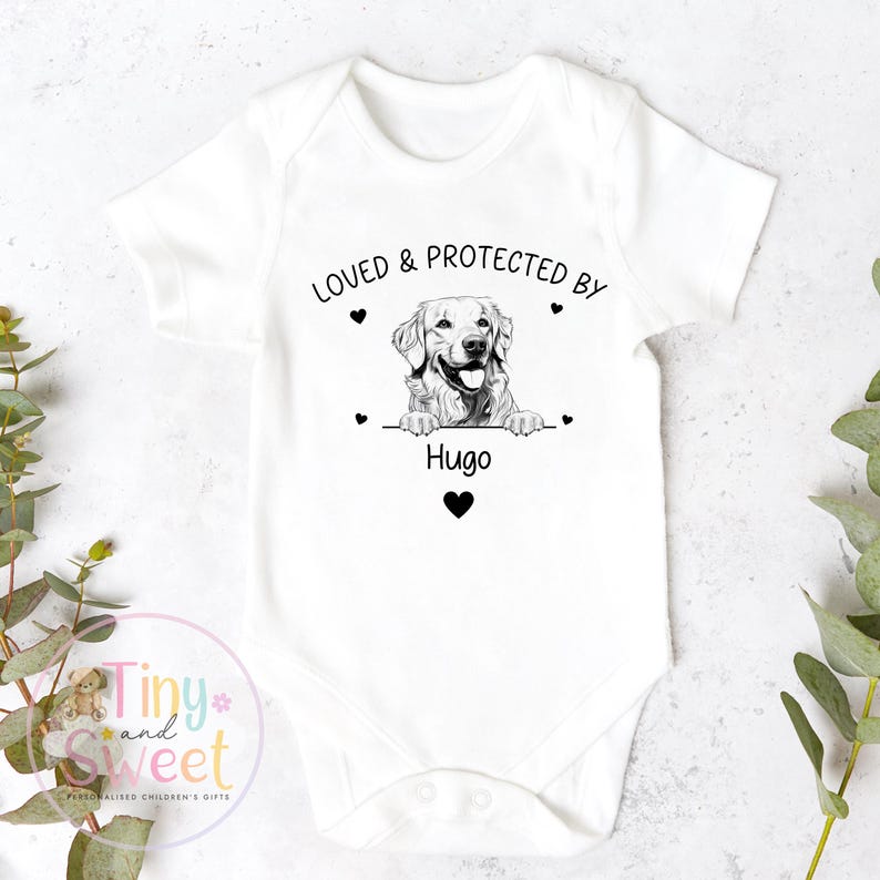Puede incluir: Un body blanco de manga corta para beb&eacute; con un estampado gr&aacute;fico en negro de un perro golden retriever apoyando sus patas sobre una l&iacute;nea. El texto "LOVED & PROTECTED BY" aparece sobre el perro y "Hugo" debajo, con peque&ntilde;os corazones.