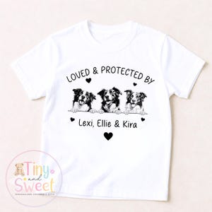 Puede incluir: Una camiseta blanca de manga corta con un gr&aacute;fico negro de tres perros asom&aacute;ndose sobre una l&iacute;nea. Sobre los perros, el texto dice LOVED & PROTECTED BY, y debajo, los nombres Lexi, Ellie & Kira, decorados con peque&ntilde;os corazones negros.