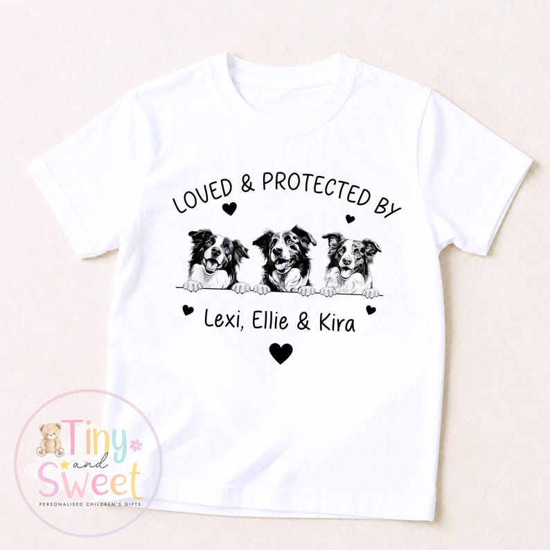 Puede incluir: Una camiseta blanca de manga corta con un gr&aacute;fico negro de tres perros asom&aacute;ndose sobre una l&iacute;nea. Sobre los perros, el texto dice LOVED & PROTECTED BY, y debajo, los nombres Lexi, Ellie & Kira, decorados con peque&ntilde;os corazones negros.