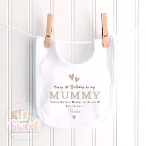 May include: A white baby bib featuring the printed text Happy 1st Birthday as my MUMMY, You're the best Mummy in the world!, Lots of love, Phoebe, accompanied by small brown heart icons. The bib is suspended by two wooden clothespins on a white string.