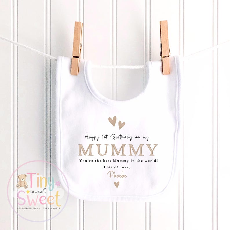 May include: A white baby bib featuring the printed text Happy 1st Birthday as my MUMMY, You're the best Mummy in the world!, Lots of love, Phoebe, accompanied by small brown heart icons. The bib is suspended by two wooden clothespins on a white string.