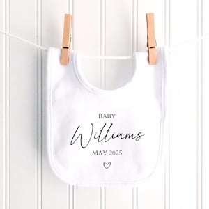 May include: A white baby bib with the name "Williams", the month "MAY 2025", and a heart symbol printed in black. The bib is hanging from a clothesline with two wooden clothespins.
