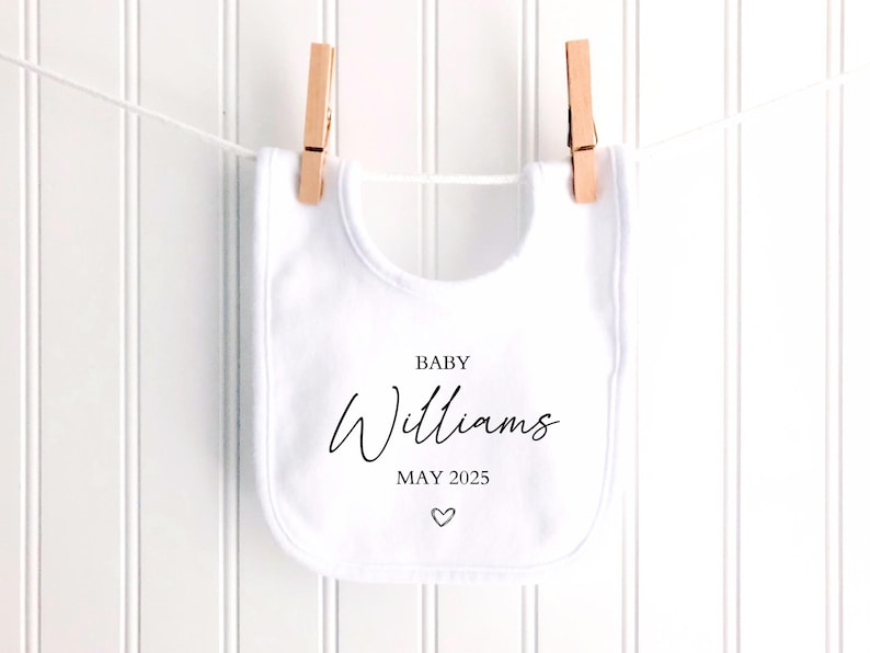 May include: A white baby bib with the name "Williams", the month "MAY 2025", and a heart symbol printed in black. The bib is hanging from a clothesline with two wooden clothespins.