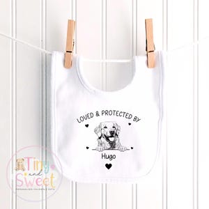 Puede incluir: Un babero blanco para beb&eacute; con un dibujo lineal negro de un perro golden retriever. El texto dice LOVED & PROTECTED BY Hugo, acompa&ntilde;ado de peque&ntilde;os iconos de corazones negros. El babero est&aacute; colgado de dos pinzas de madera en una cuerda.