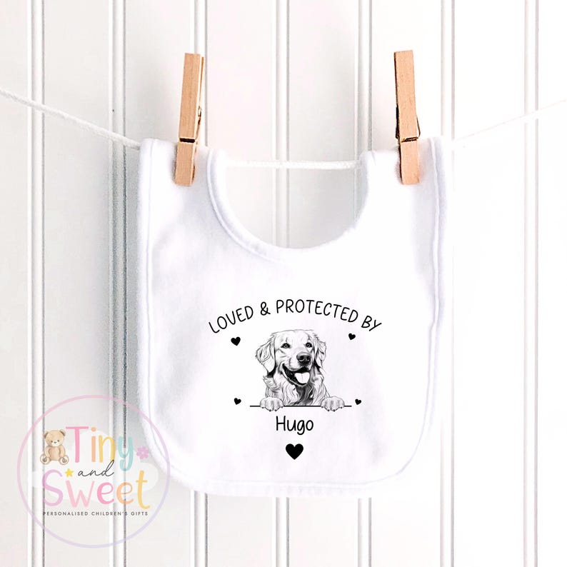 Puede incluir: Un babero blanco para beb&eacute; con un dibujo lineal negro de un perro golden retriever. El texto dice LOVED & PROTECTED BY Hugo, acompa&ntilde;ado de peque&ntilde;os iconos de corazones negros. El babero est&aacute; colgado de dos pinzas de madera en una cuerda.