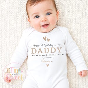 May include: A white long-sleeved baby bodysuit featuring the printed text Happy 1st Birthday as my DADDY, You're the best Daddy in the world!, Love from, Ronnie x, decorated with small brown heart icons above and below the text.