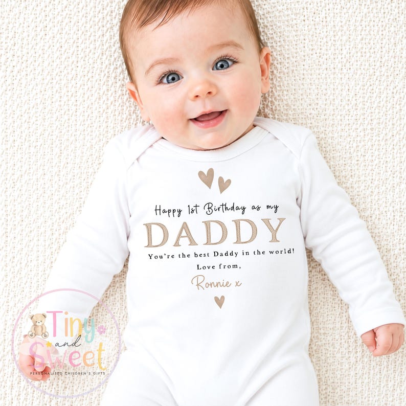 May include: A white long-sleeved baby bodysuit featuring the printed text Happy 1st Birthday as my DADDY, You're the best Daddy in the world!, Love from, Ronnie x, decorated with small brown heart icons above and below the text.