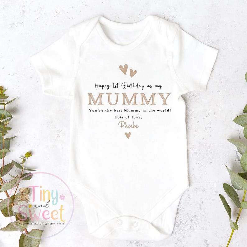 May include: A white short-sleeved baby bodysuit featuring the text Happy 1st Birthday as my MUMMY, You're the best Mummy in the world! Lots of love, Phoebe, accented with small tan heart icons printed in a centered, clean, and legible font style.