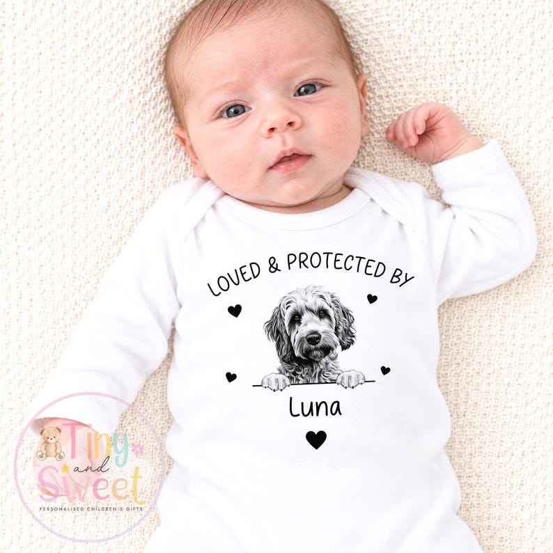 Puede incluir: Un body blanco de manga larga para beb&eacute; con un gr&aacute;fico negro de un perro apoyando sus patas sobre una l&iacute;nea. Sobre el perro, el texto dice LOVED & PROTECTED BY, y debajo del perro, el nombre Luna est&aacute; impreso junto a peque&ntilde;os corazones negros.
