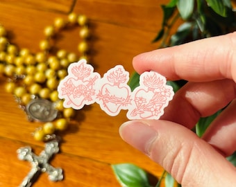 May include: A small sticker featuring three distinct heart illustrations outlined in light pink. The left heart is pierced by a sword, the center heart is encircled by a crown of thorns with a cross on top, and the right heart is adorned with flowers.