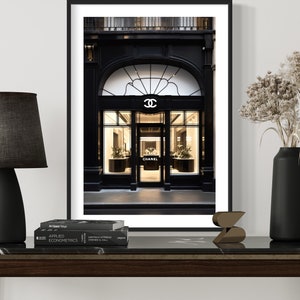 May include: A framed print of a Chanel store facade with a black and white color scheme. A black lamp with a gray shade, a black vase with dried flowers, and a decorative object sit on a dark wood table.