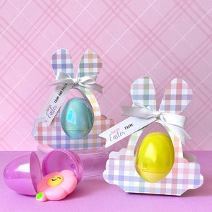 Puede incluir: Dos soportes para huevos de Pascua en forma de conejo de cuadros pastel con lazos de cinta blanca. Cada soporte tiene un huevo de plástico transparente en su interior. Un huevo es azul iridiscente y el otro es amarillo. Los soportes tienen etiquetas que dicen "Feliz Pascua" y "De".