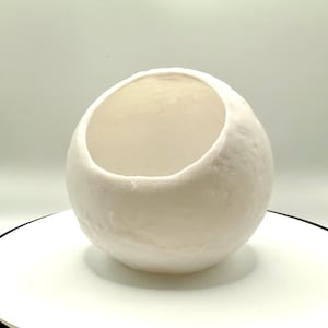 May include: A white, egg-shaped ceramic bowl with a textured surface. The bowl has a large opening at the top and is designed to hold items. The bowl is a neutral color and could be used for decorative or functional purposes.
